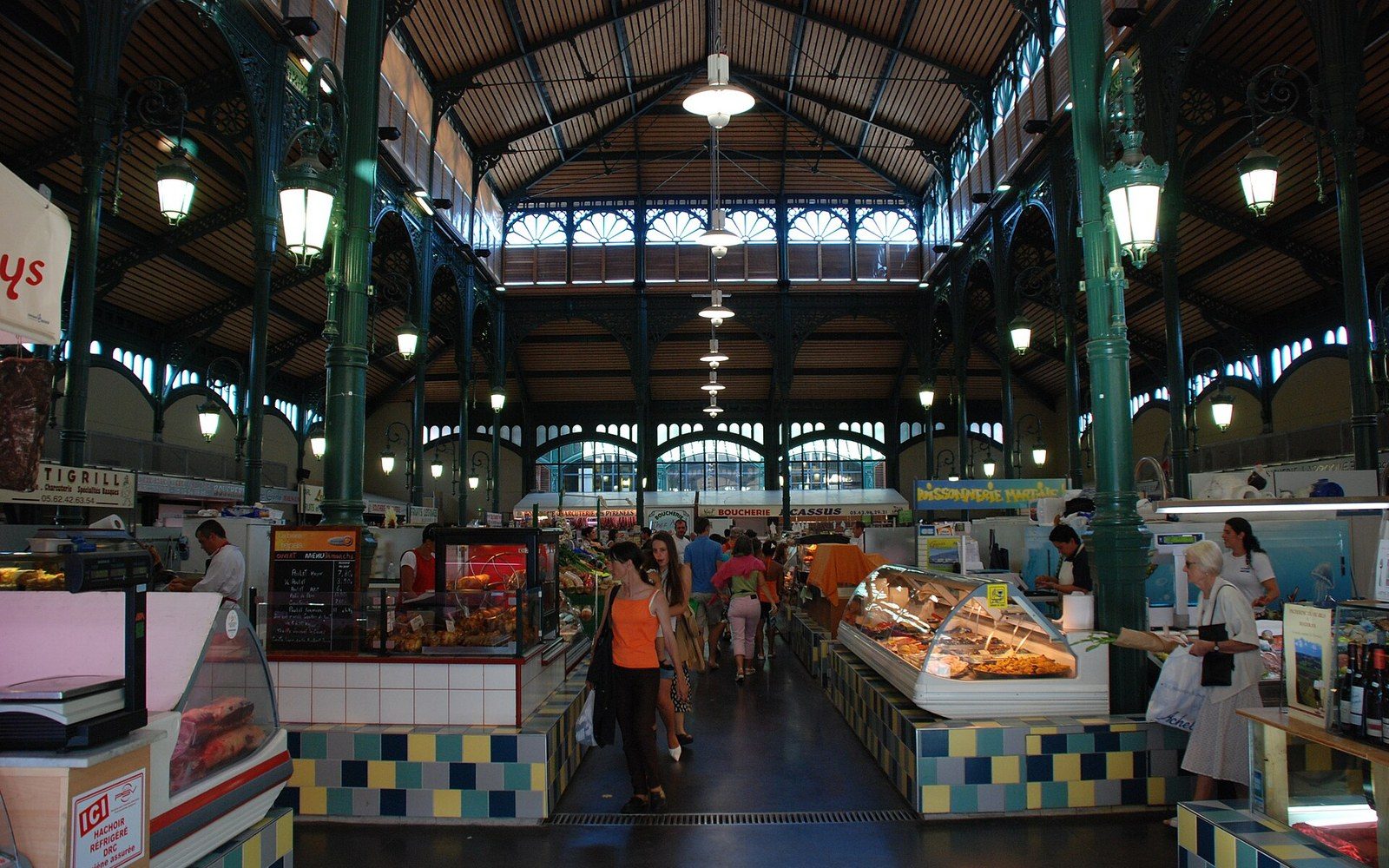 A covered market hall with iron columns, stalls stacked with produce, adults shopping